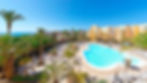 Bright resort scene with a large pool, surrounded by palm trees and sun loungers, yellow buildings, and a clear blue sky, overlooking the sea.