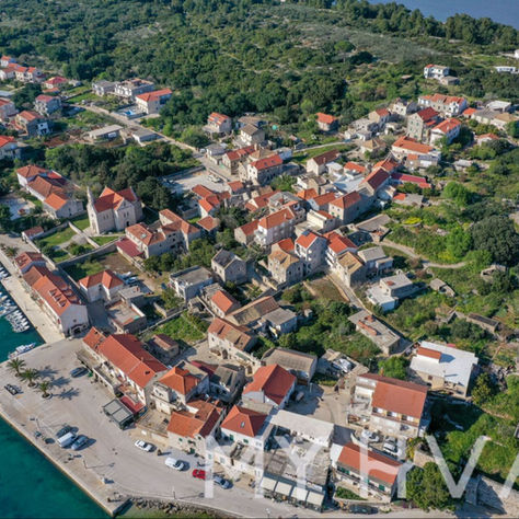 Aerial view of a seaside village with red-roofed houses, a small harbor full of boats, and green hills by blue water.
