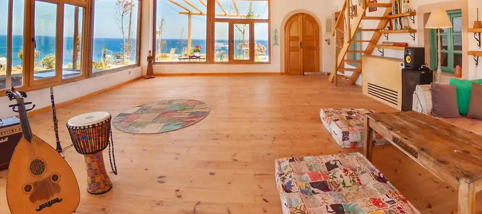 Sunlit room with wooden floors, patchwork cushions, a drum, lute, and clarinet. Ocean view through large windows. Cozy, artistic vibe.
