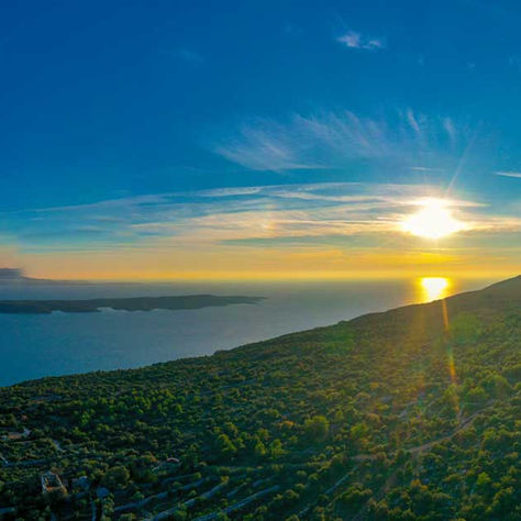 Sunset over a green coastal hillside and calm blue sea, with an island on the horizon and golden light on the water.