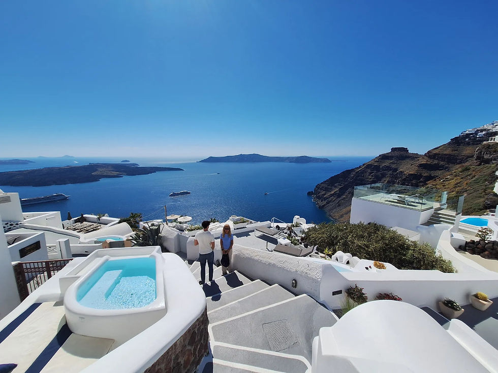 Two people walk down white stone steps beside a pool, overlooking a bright blue sea and mountainous coastline under a clear sky.