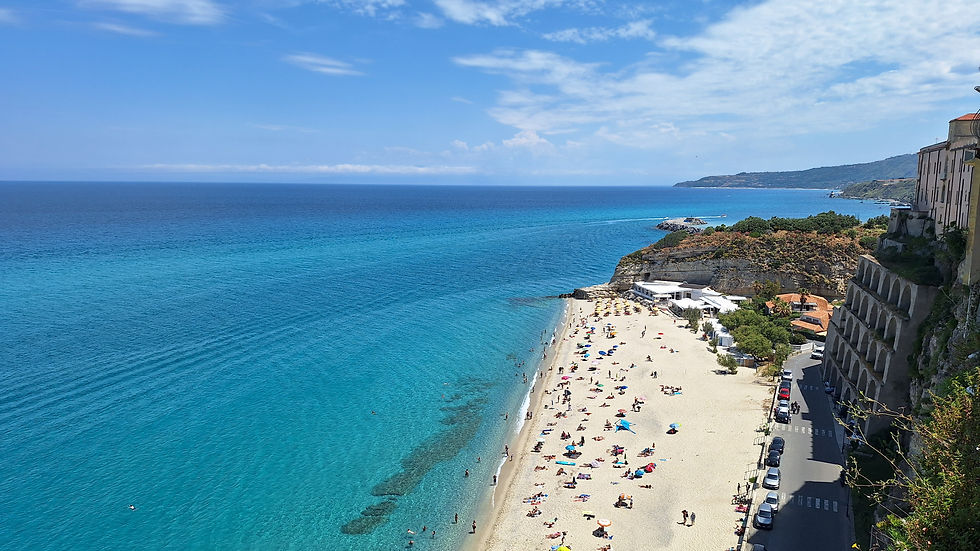 Sunny aerial view of a crowded turquoise beach beside cliffs and buildings, with swimmers, umbrellas, and calm blue sea.