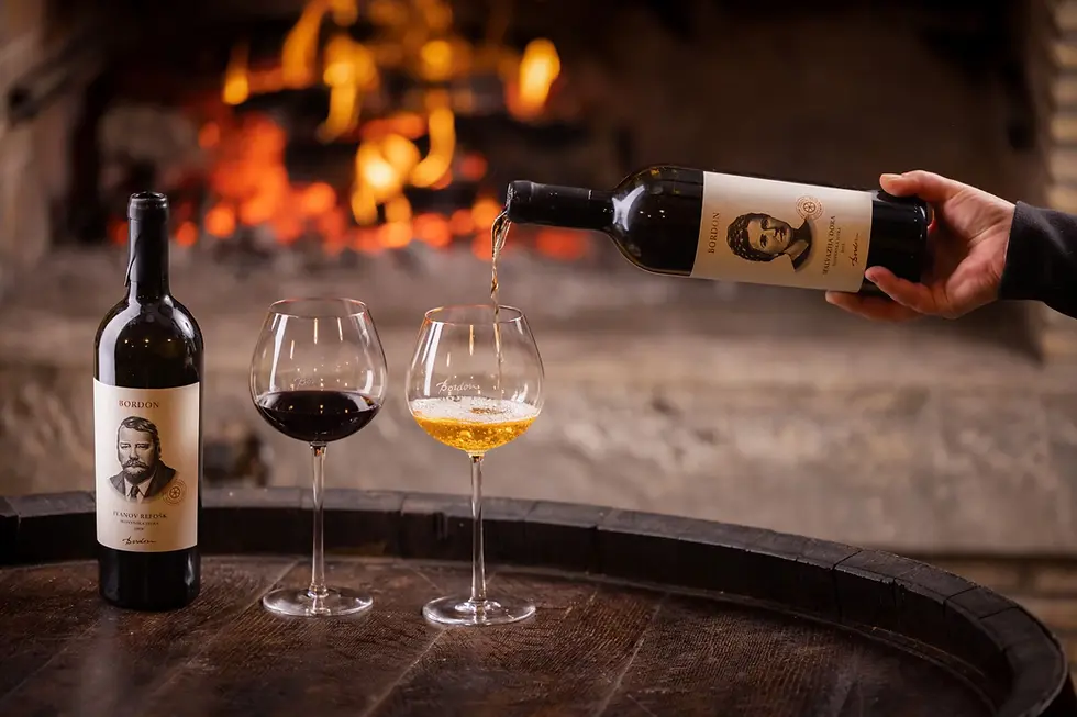 Hand pours Bordon red wine into a glass beside another bottle and glass, with a glowing fireplace in the background.