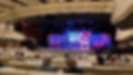 Audience seated in a theater watching a stage with a colorful "Buddy's Diner" set. The ceiling has rows of spotlights and elegant decor.