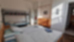 Sunlit bedroom with a double bed and bunk bed. Blue and gray bedding, wooden dresser, hangers, and striped shower curtain in the background.