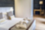 Modern bedroom with a neatly made bed featuring "Boston" pillows, folded robes, and towels. Elegant lamp and desk on wooden floor. Cozy mood.