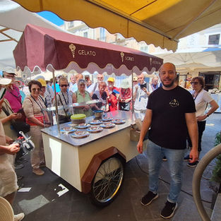 Crowd gathers around a gelato cart under awnings in a sunny plaza, with GELATO, SORBET and ICE CREAM on the maroon canopy.
