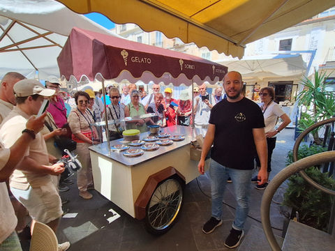 Crowd gathers around a gelato cart under awnings in a sunny plaza, with GELATO, SORBET and ICE CREAM on the maroon canopy.
