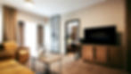 Hotel room with mustard sofa, wall art of a stork, and a TV displaying "Terme Vivat." Bright, modern decor with warm tones.