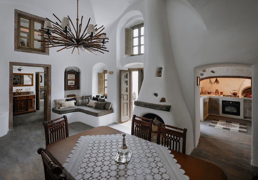 Cozy living room with arched fireplace, rustic chandelier, and dining table. Cushioned bench seat by wood-framed windows, open doorway to kitchen.