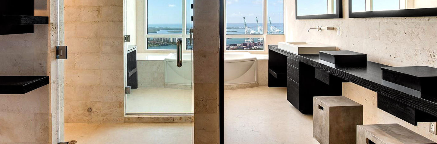Miami Beach master bathroom renovation featuring natural stone walls, marble flooring, custom oak wood vanity, and built-in tub with panoramic ocean views.