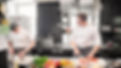Two chefs working together to prepare dishes in a busy commercial kitchen filled with cooking equipment and ingredients.