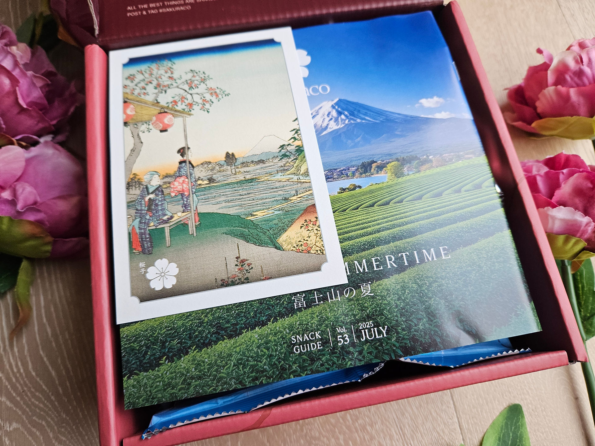 Opening the Sakuraco subscription box packaging, surrounded by pink flowers. Postcard and 24 page mini magazine visible. LisaEatsTheWorld.com