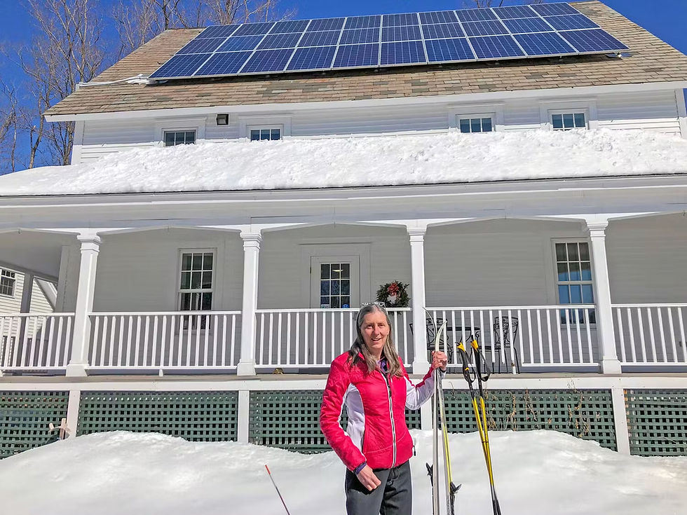 Kim Quirk, CPCNH Board Chair, standing in front of a snow-covered white home with rooftop solar panels and cross-country skis in hand.