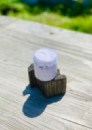 Lavish Lavender Whipped Body Butter colored light purple on top of dark brown wood piece sitting on top of a rustic grey table with green grass in the background outside.