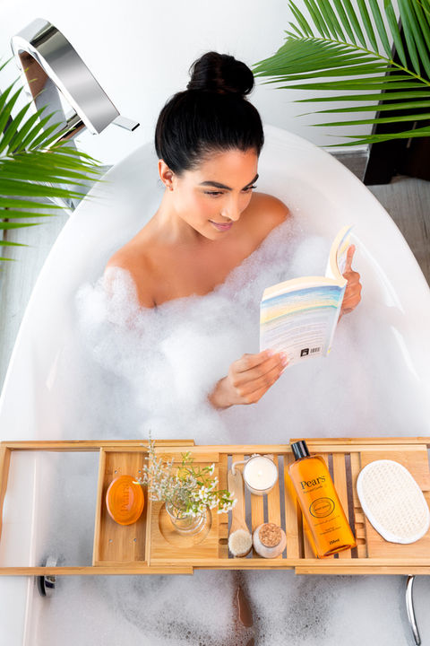 A woman relaxing in a bubble bath with a book, surrounded by Pears body wash, Pears soap, and spa essentials on a bamboo bath tray. Luxury self-care and skincare photography.
