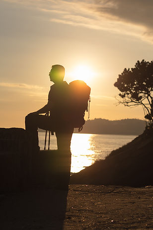 homem em pé com mochilão nas costas e perna apoiada em uma mureta apreciando a vista e ao fundo o por do sol no mar 