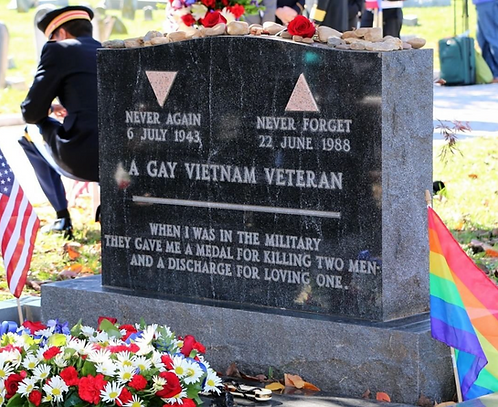 The tombstone of gay American Vietnam veteran, reading the words: "When I was in the military, they gave me a medal for killing two men, and a discharge for loving one." The tombstone is made with reflective black granite and contains two pink triangles, referencing the symbol used to mark gays in nazi concentration camps. An American flag on one side of the tombstone, and a pride flag on the other side. Flowers decorate the front of the tombstone.