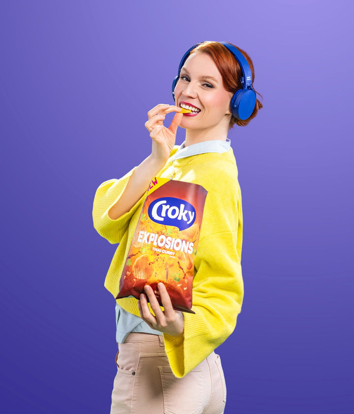 Een roodharige dame tegen een paarse achtergrond en gele trui, blauw koptelefoon en een zak chips.