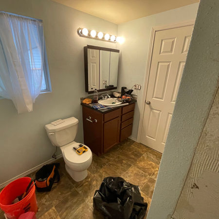 A bathroom before remodeling, displaying a toilet, sink, and mirror, with dated tiles and minimal furnishings.