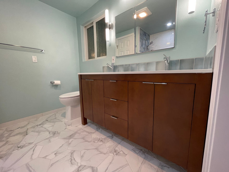 Modern bathroom featuring a wood vanity with quartz counter and aqua tile backsplash, pale teal walls, a marble-patterned tile floor, and a window for natural light.