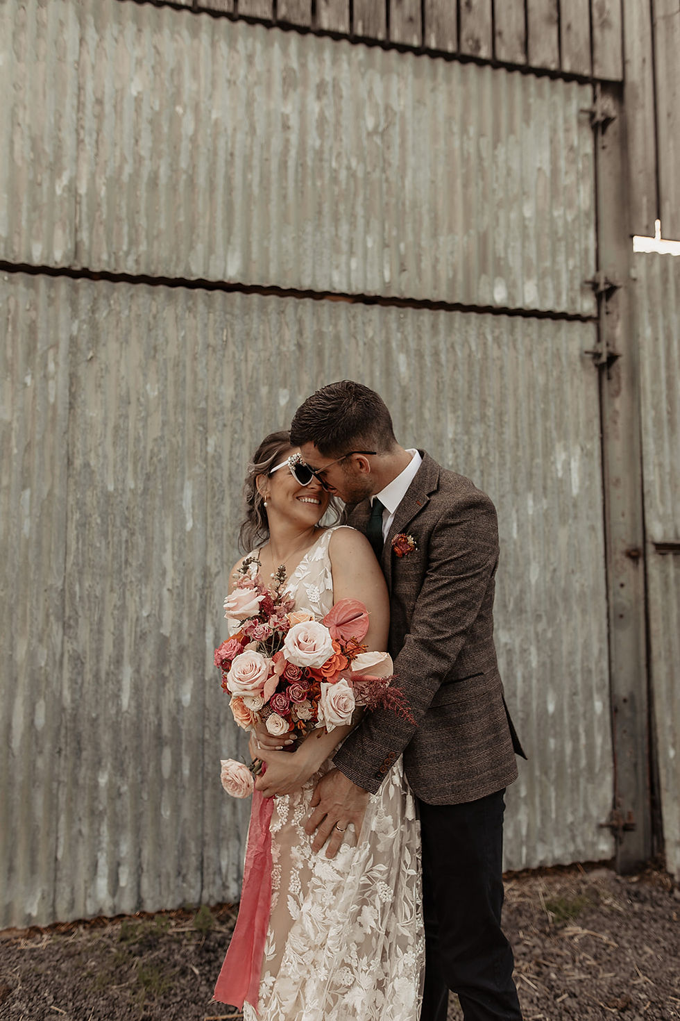 Couple smiling, embracing near corrugated metal wall. Woman holds vibrant bouquet, wearing sunglasses; joyful and intimate moment captured.