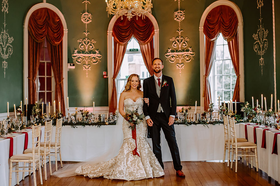 Bride in lace gown and groom in suit stand smiling in elegant dining room with draped windows, chandeliers, and candlelit tables.