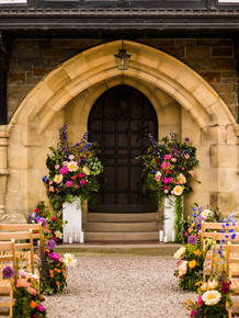 Wedding ceremony set up in front of an arched doorway with large colourful floral displays on white plinths