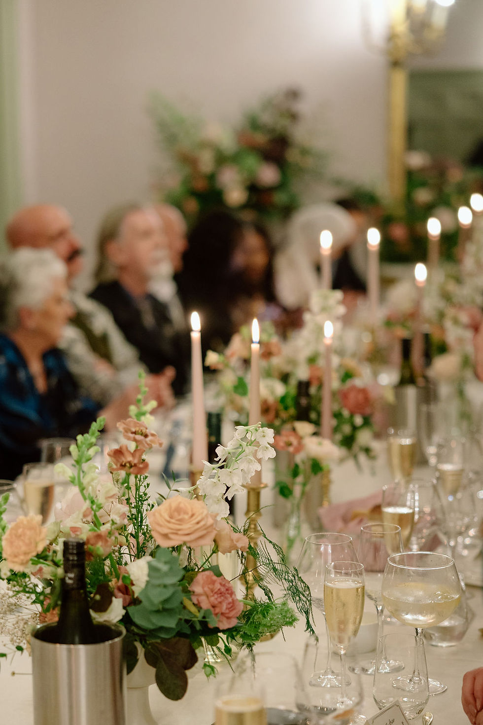 Elegant dinner table with floral centerpieces and lit candles. Guests sit blurred in the background, creating a warm, festive atmosphere.