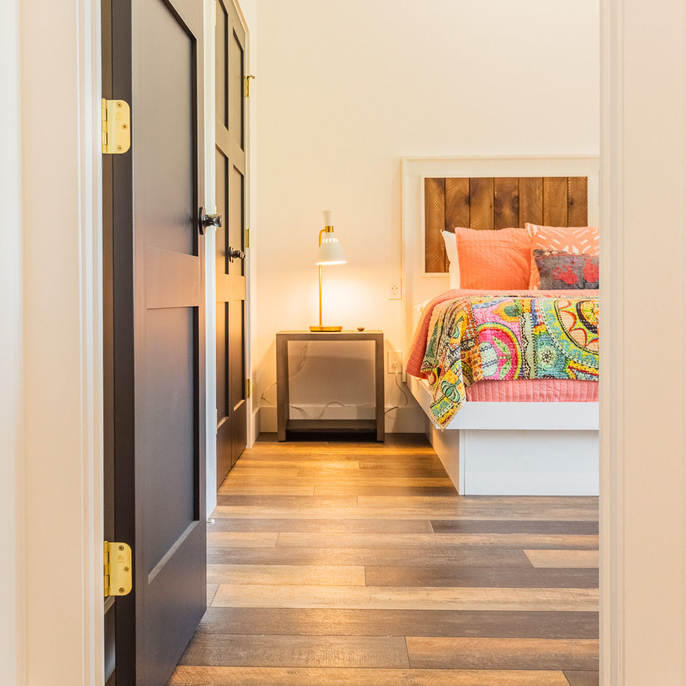 A warm and inviting bedroom featuring a wooden-accented bed frame, vibrant bedding, and a sleek bedside lamp, complemented by dark-paneled doors and stylish flooring.