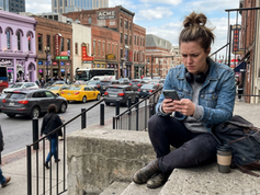 Woman in denim jacket, headphones around neck, sitting on city steps using smartphone. Busy street with traffic, buildings, vibrant signs.