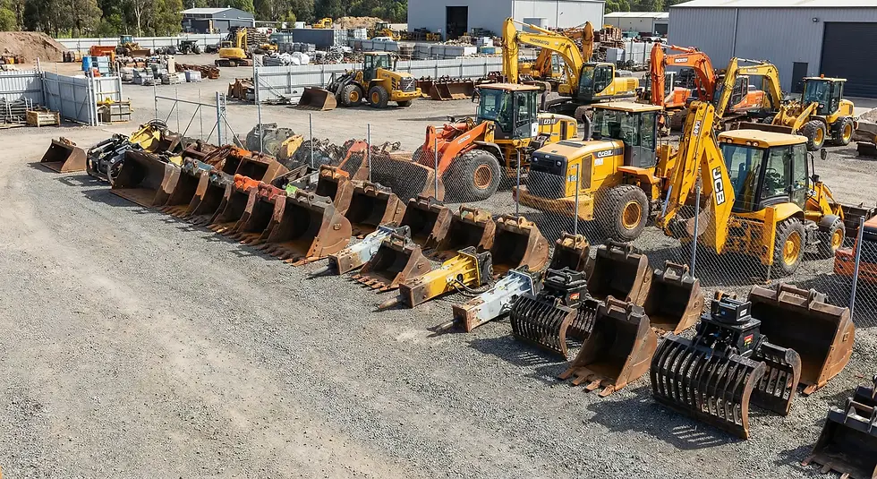 Construction yard with yellow and orange excavators and backhoes. Various large metal buckets lined up. Industrial setting with fence.