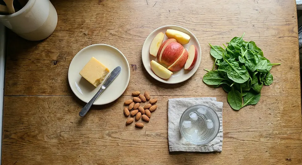 Cheese with knife, apple slices, almonds, spinach, and a glass of ice water on a wooden table create a fresh, balanced snack scene.