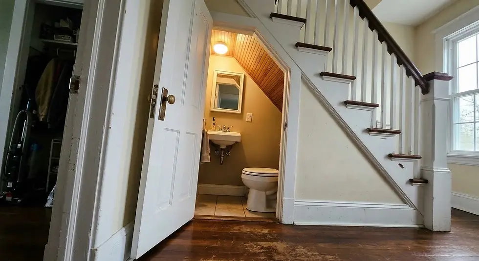 A small bathroom with a sink and toilet is built under a wooden staircase. An open closet is visible on the left, with coats and a vacuum.