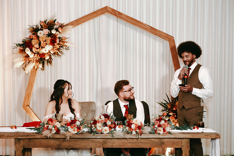Bride and groom seated at a table with floral decor. A man in a suit stands, speaking into a microphone. Warm, festive atmosphere.
