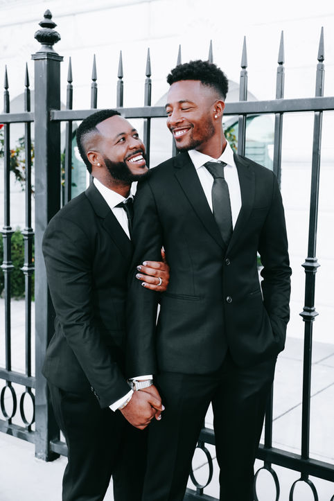Two grooms smiling at each other while posing in black suits outside the Clark County Government Center in downtown Las Vegas.