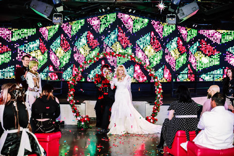 Two brides celebrating during a vibrant anime inspired wedding ceremony in front of a colorful LED wall at Substance nightclub on Fremont Street in Las Vegas.