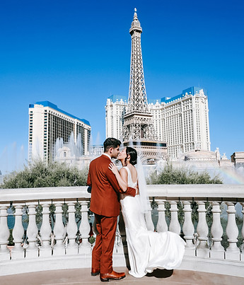 Non traditional couple kiss with the Bellagio fountains and the Paris hotel in the background as the wind blows the bottom of her gown
