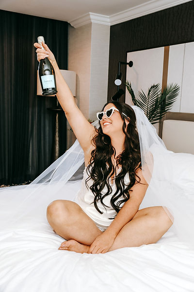 Bride sits crosslegged on her l bed raising a champagne bottle with heart shaped sunglasses, silk pajamas, an a cathedral veil