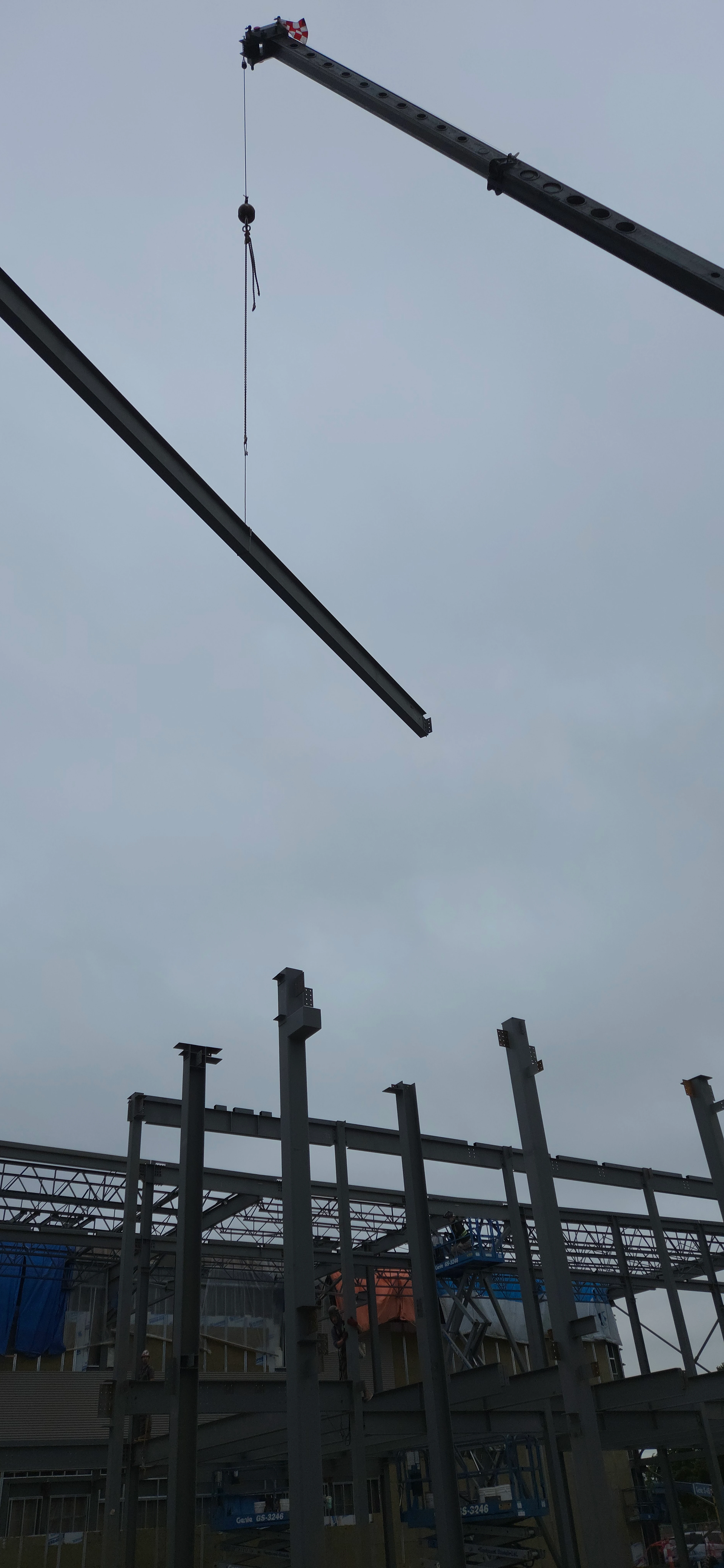 A low-angle shot looking up as a crane lowers a long steel beam towards the steel skeleton of a building under construction, all against an overcast sky.