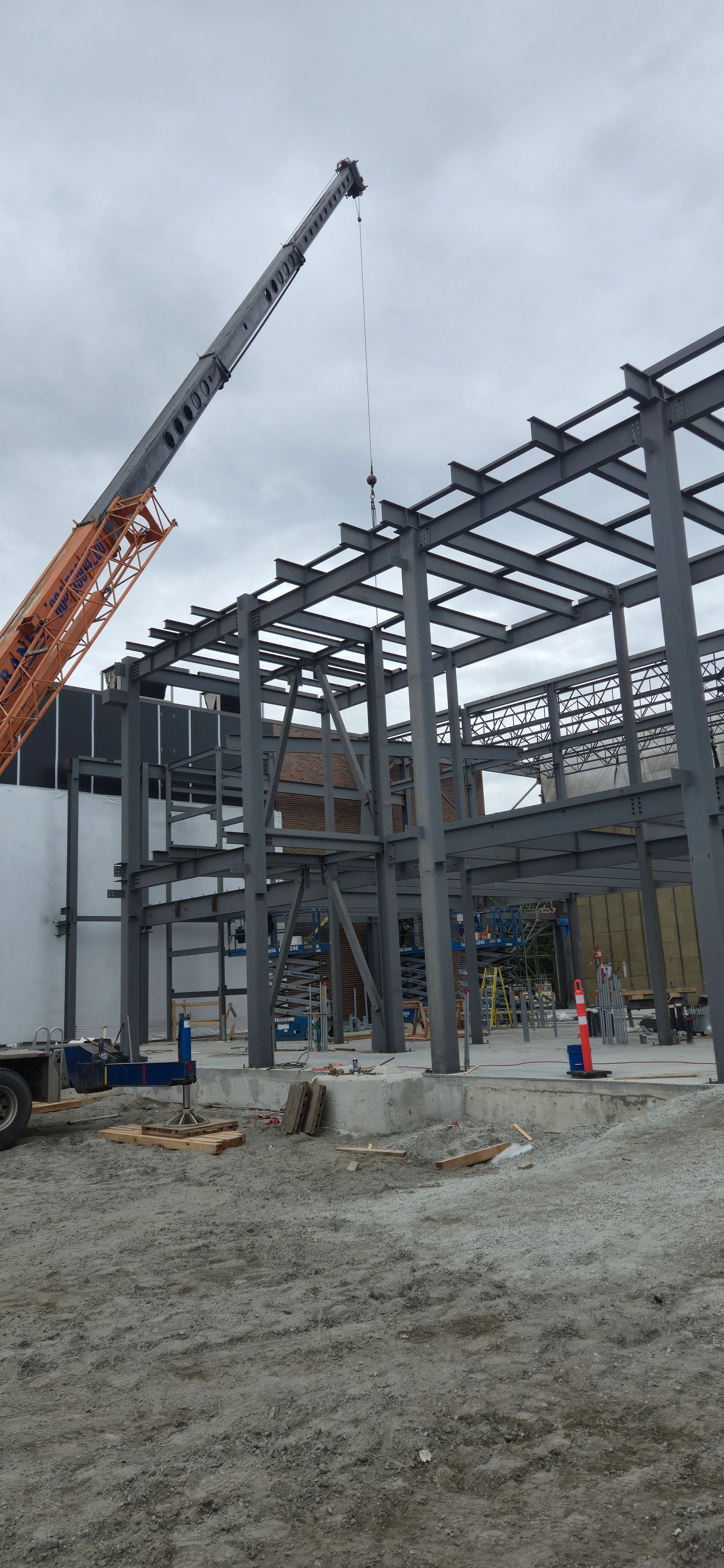 A wide-angle view of a multi-story steel building frame under construction, with an orange and grey mobile crane arm extending over it from the left.