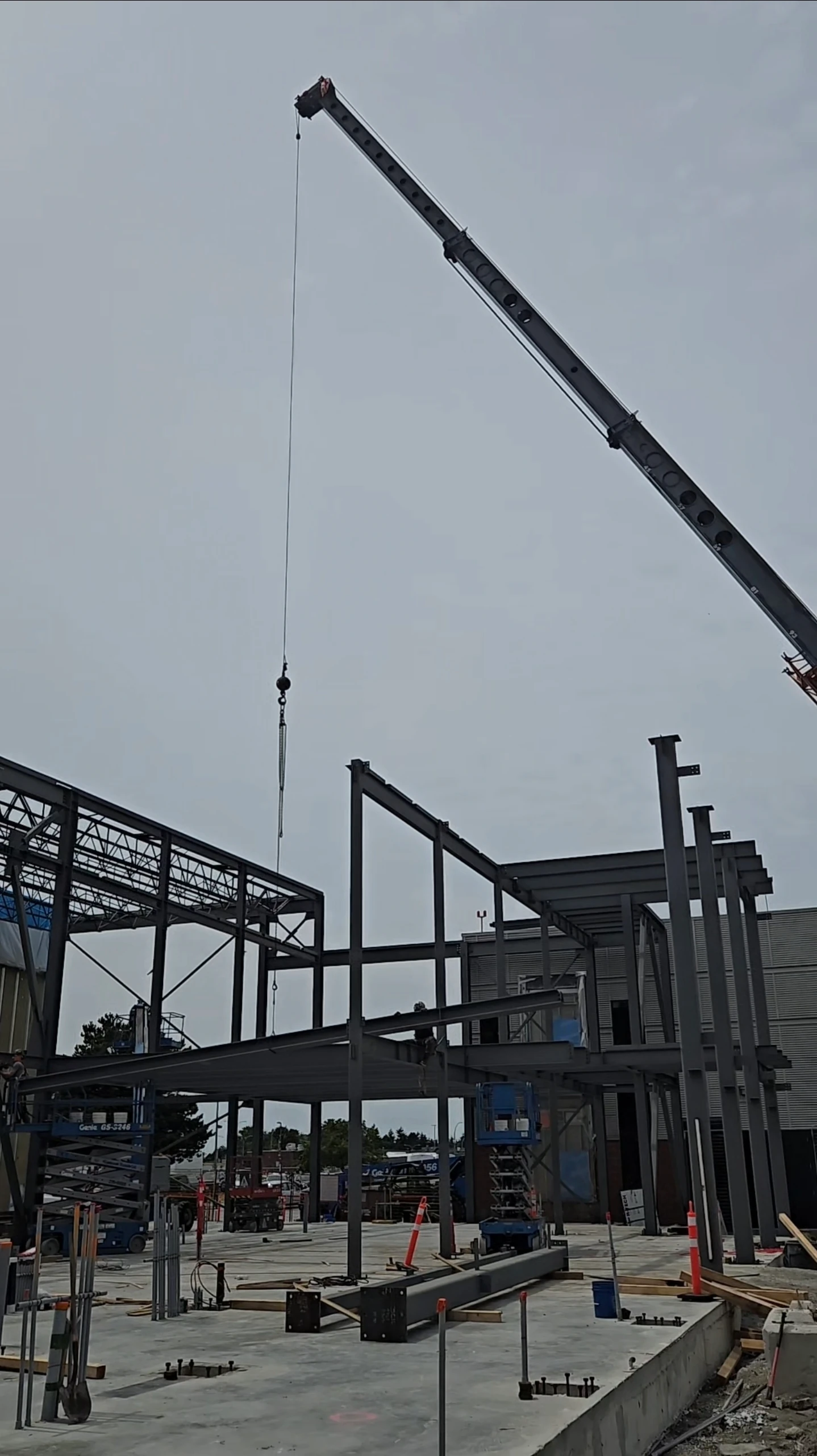 A construction site with the steel framework of a building being erected against an overcast sky. A large crane arm extends from the top right, lowering its hook and cable toward the structure.