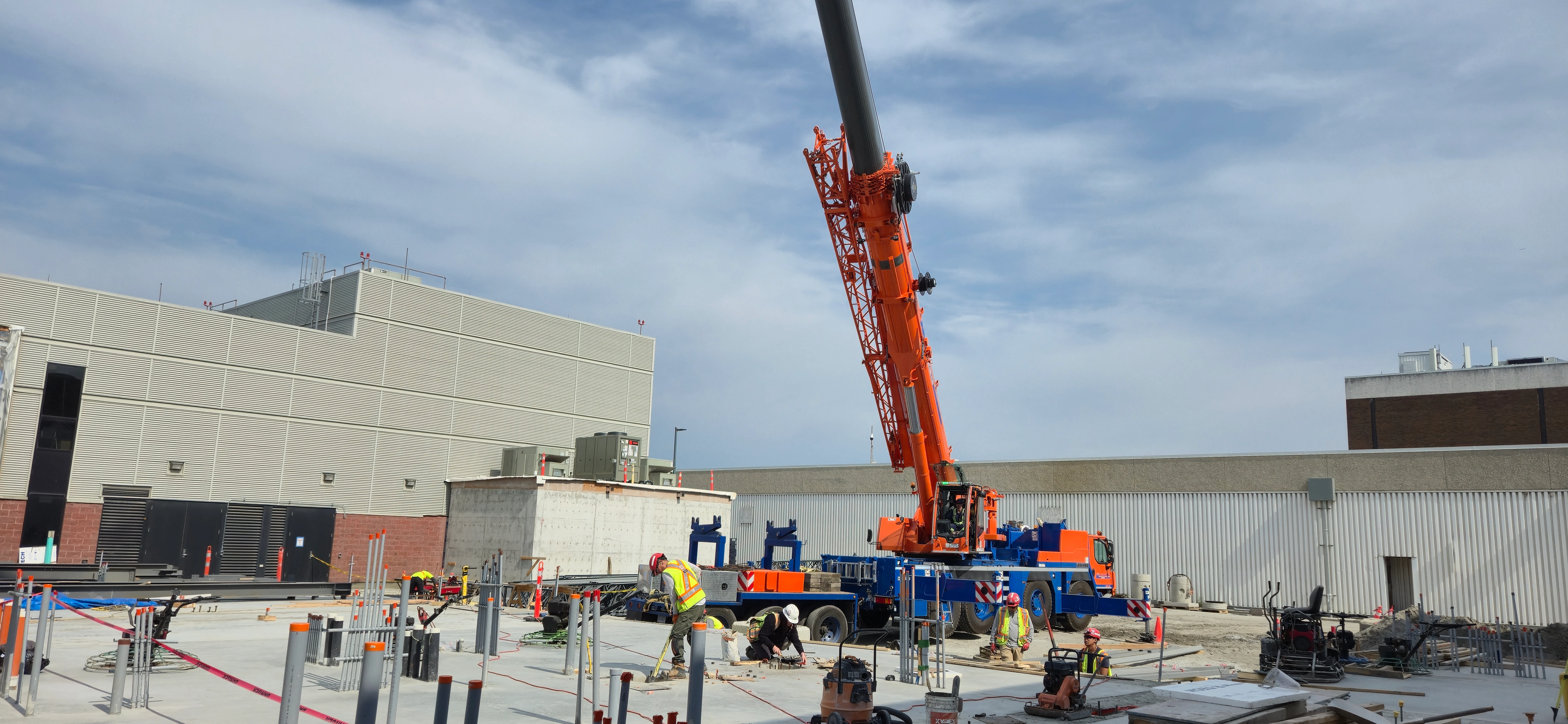 A wide shot of a construction site with a large, orange and blue mobile crane as the central focus. Several workers in safety vests are on the concrete foundation around the crane, with large buildings in the background.