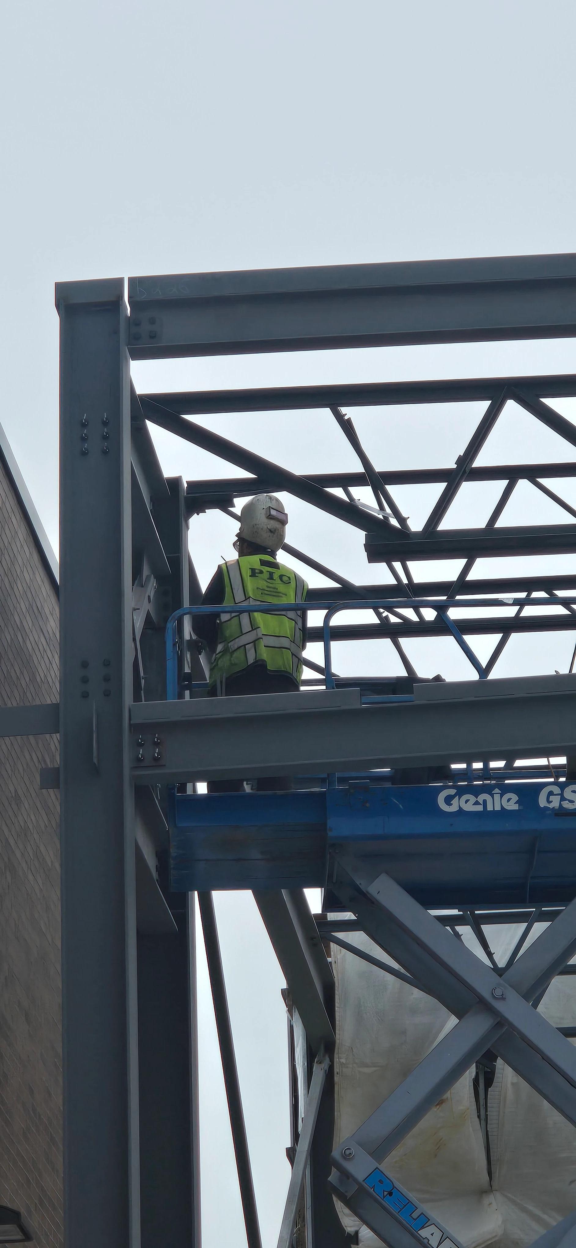 A construction worker in a yellow safety vest and hard hat is visible on an elevated platform, positioned within the steel framework of a building under construction.