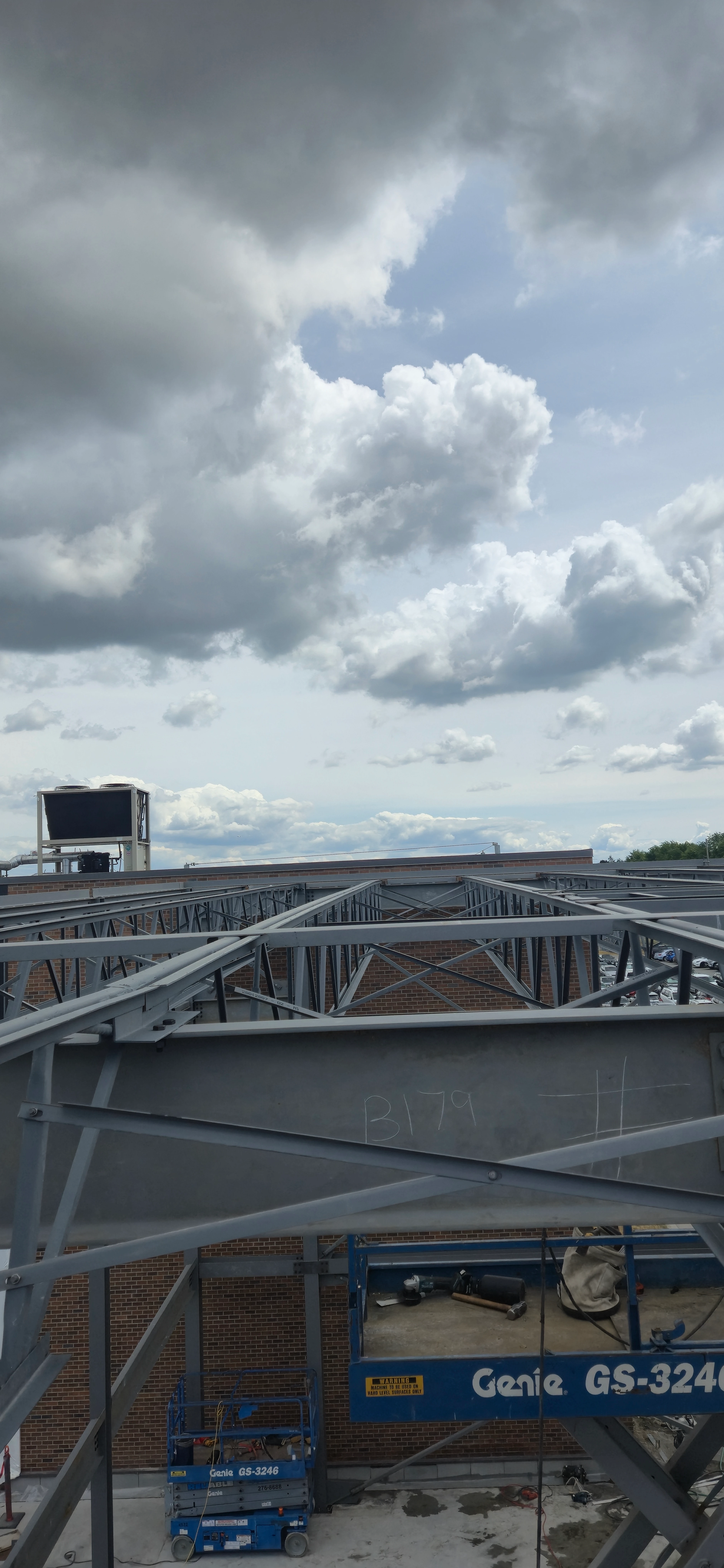 A high-angle view looking across the steel roof joists of a building under construction. A blue scissor lift is visible on the concrete floor below, next to a brick wall. A large rooftop HVAC unit is in the background.