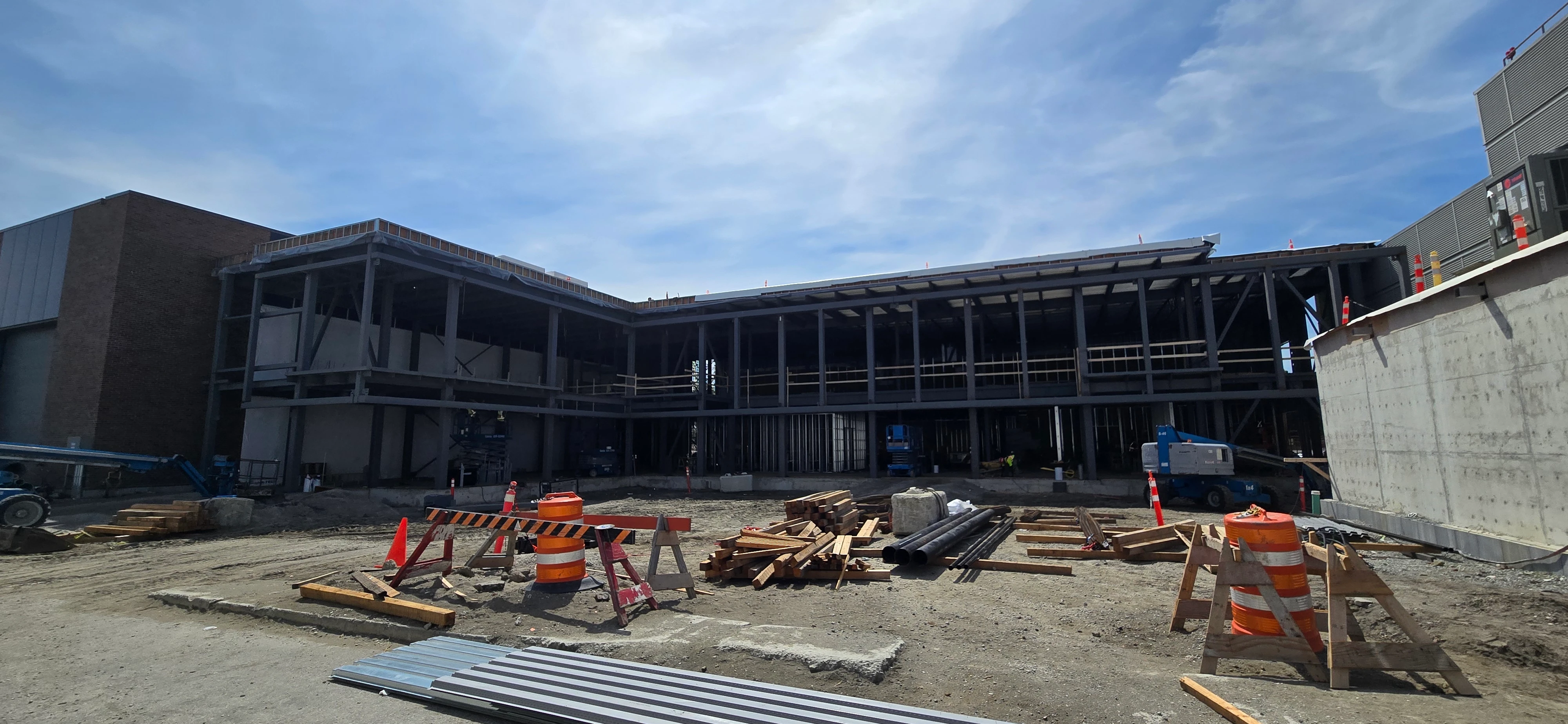 A wide shot of a construction site on a sunny day, showing the steel framework of a large building in progress. Various construction materials, barriers, and equipment are scattered in the foreground.