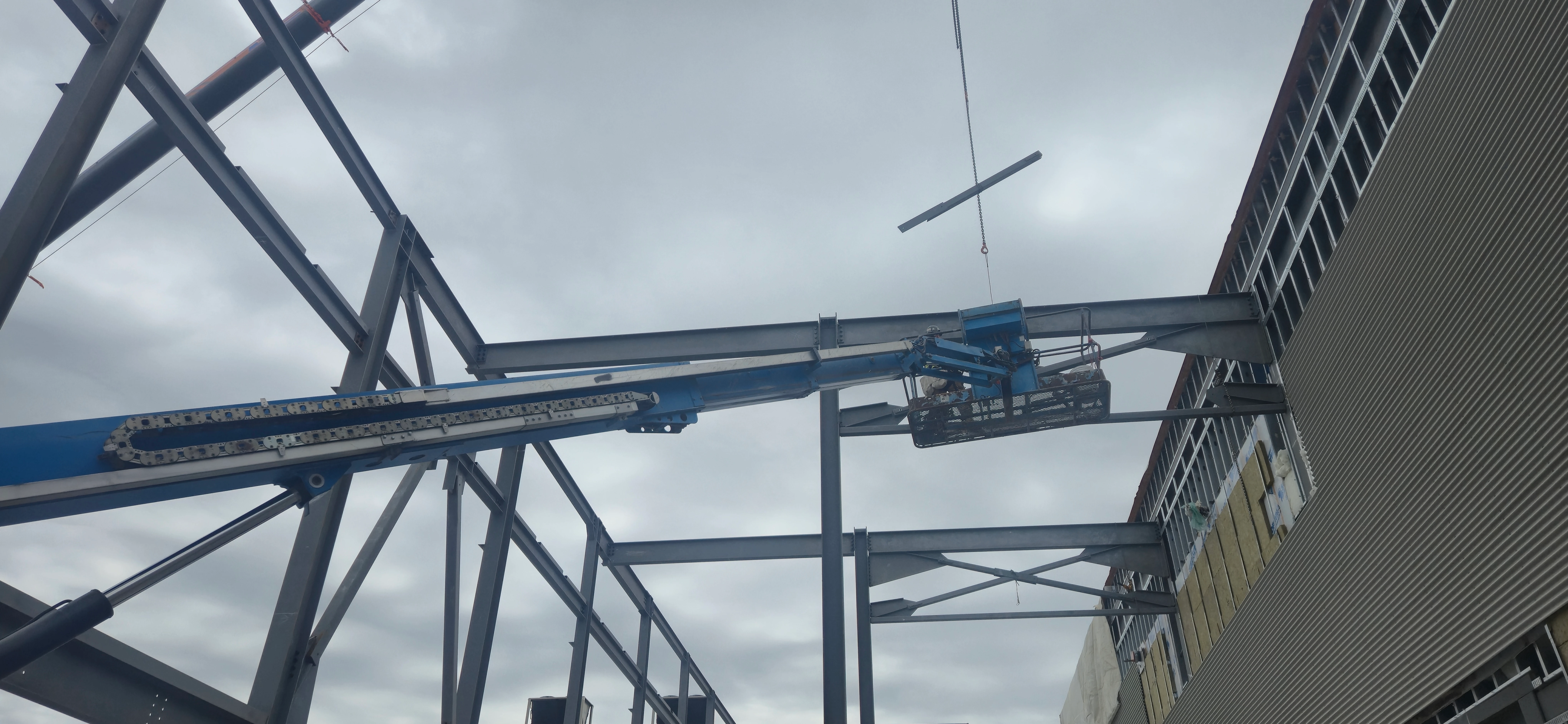 A construction worker in a blue boom lift basket is positioned high up, working on the steel frame of a building. A crane is lowering a small steel piece on a cable from above.