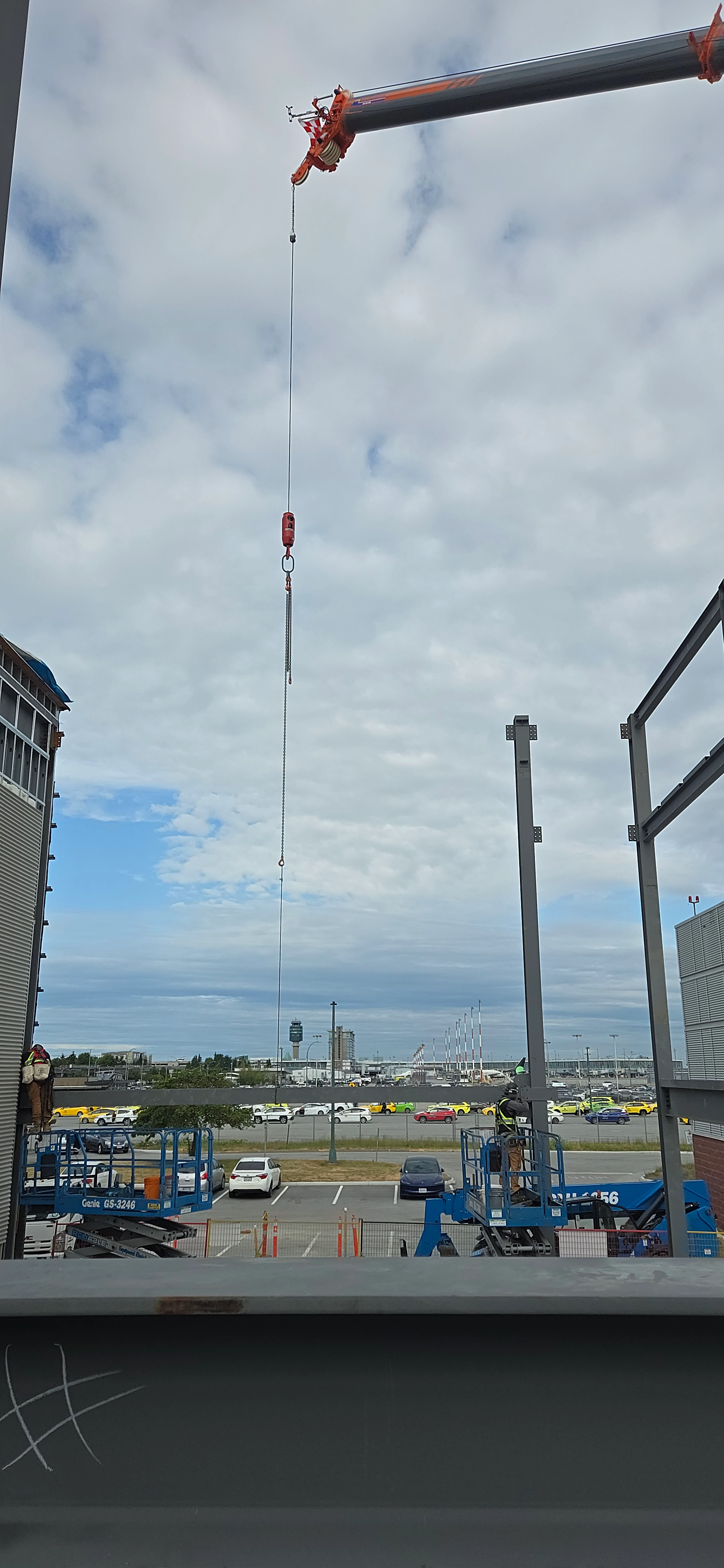 A high-angle view from a construction site, looking down on a parking lot. A crane arm extends from the top of the frame with its hook and cable hanging down. An airport control tower is visible in the background.
