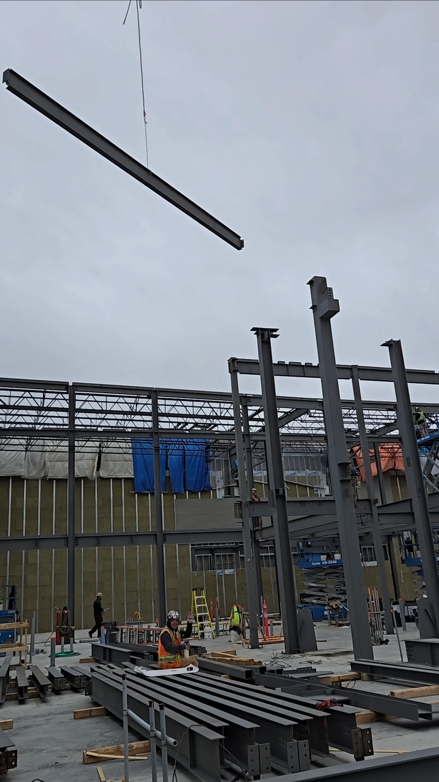 A construction site with a large metal beam being lowered by a crane. Workers in safety vests and hard hats are visible below, and the framework of a building is in the background.