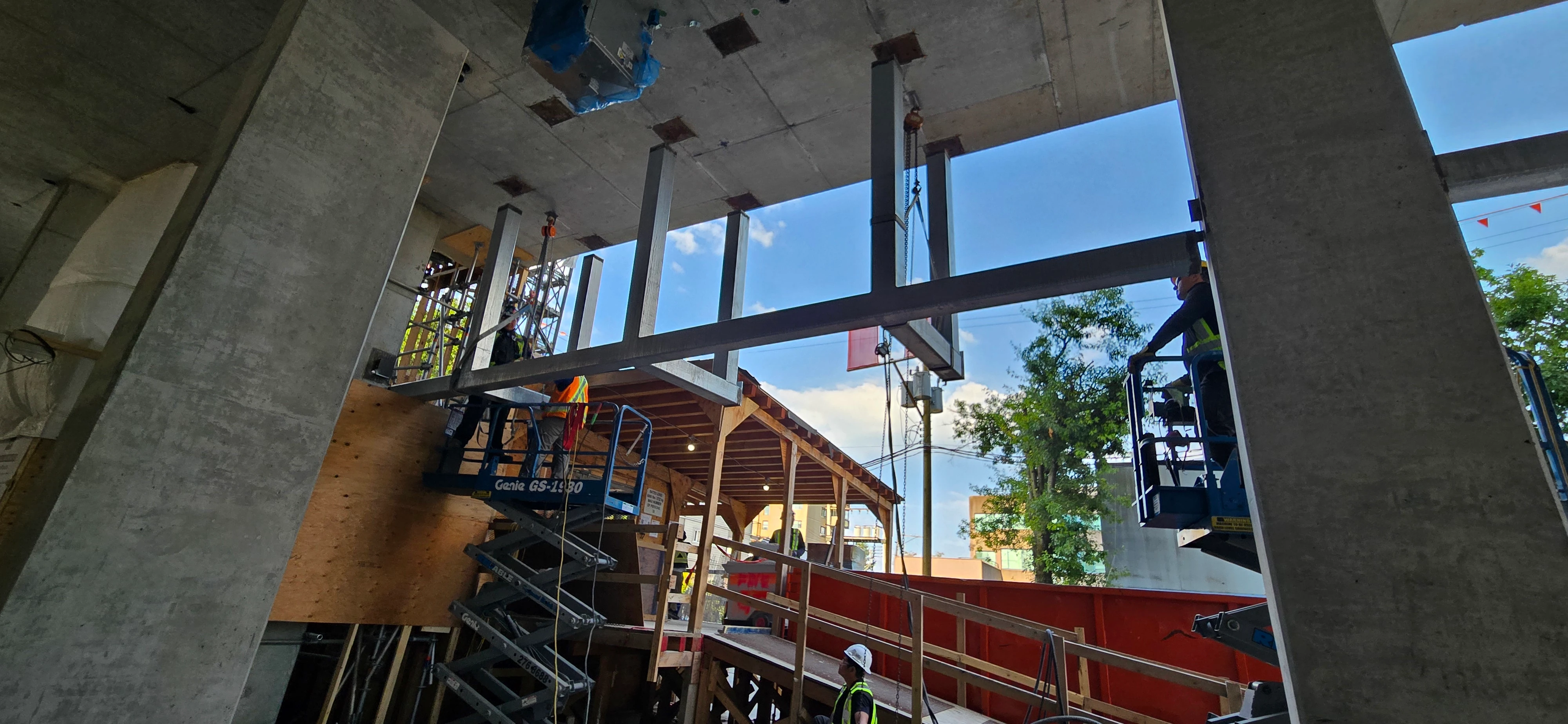 Construction site view from below, showing workers on scissor lifts installing large horizontal steel I-beams into a concrete ceiling. Concrete pillars frame the scene, and outdoor greenery is visible in the background.